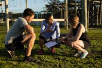 Front view of a young Caucasian woman and a young Caucasian man squatting down with a young Caucasian female trainer holding a clipboard and talking at an outdoor gym during a bootcamp training session — Stock Photo