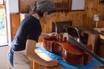 Seitenansicht einer älteren kaukasischen Geigenbauerin, die an einem Cello auf einer Werkbank in ihrer Werkstatt arbeitet, im Hintergrund hängen Werkzeuge an der Wand — Stockfoto