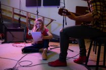 Передній вид на молоду кавказьку жінку, що сидить на підлозі і співає з мікрофоном і тримає текст, в той час як молодий змішаний чоловік, що сидить на стільці, грає на акустичній гітарі. — стокове фото