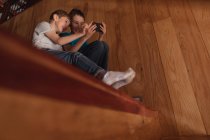 Vista lateral de cerca de dos niños preadolescentes sentados en una escalera en casa, utilizando un teléfono inteligente - foto de stock