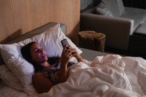 Vue élevée d'une jeune femme brune caucasienne couchée sur le dos au lit à l'aide d'un smartphone — Photo de stock