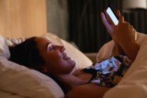 Вид збоку крупним планом усміхнений молодий Кавказький жінка брюнетка лежачи на спині в ліжку за допомогою смартфона — стокове фото