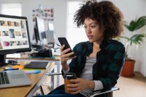 Вид збоку крупним планом молода змішана жінка, що сидить за столом і використовує смартфон у творчому офісі — стокове фото