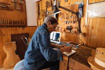 Side view of a senior Caucasian female luthier making notes in her workshop, with a laptop and a tablet computer in front of her and tools hanging up on the wall in the background — Stock Photo