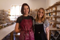 Portrait of two young Caucasian female potters wearing aprons standing, embracing and looking to camera smiling in a pottery studio — Stock Photo