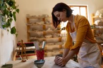 Vista lateral de uma jovem oleiro caucasiana em pé em uma mesa de trabalho e amassar um pedaço de argila em um estúdio de cerâmica — Fotografia de Stock