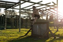 Seitenansicht eines hemdlosen jungen kaukasischen Mannes, der während eines Bootcamp-Trainings in einem Outdoor-Fitnessstudio über eine Mauer springt — Stockfoto