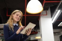 Nahaufnahme einer jungen kaukasischen Frau, die mit einem Tablet-Computer im modernen Büro eines kreativen Unternehmens vor der Kamera steht und lächelt — Stockfoto