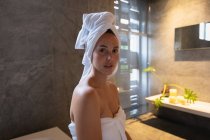 Retrato de uma jovem mulher morena caucasiana vestindo uma toalha de banho e com o cabelo envolto em uma toalha, olhando diretamente para a câmera em um banheiro moderno — Fotografia de Stock