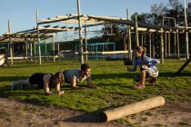 Side view of a young Caucasian woman and a young Caucasian man doing the plank exercise while a young Caucasian woman times them at an outdoor gym during a bootcamp training session — Stock Photo