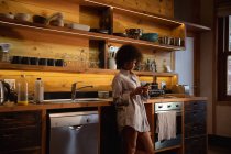 Side view of a young mixed race woman wearing a shirt standing using a smartphone at home in her kitchen — Stock Photo