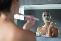 Sobre a vista do ombro de uma jovem mulher morena caucasiana vestindo uma toalha de banho olhando no espelho segurando um frasco e aplicando um pacote de rosto com uma escova em um banheiro moderno — Fotografia de Stock