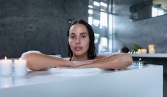 Portrait of a young Caucasian brunette woman sitting in a foam bath with lit candles on the edge, leaning on the side and looking straight to camera — Stock Photo