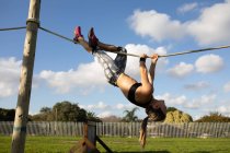 Vista laterale di una giovane donna caucasica appesa a una corda orizzontale su un telaio da arrampicata in una palestra all'aperto durante una sessione di allenamento bootcamp — Foto stock