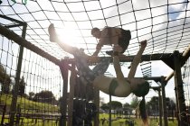 Seitenansicht einer jungen kaukasischen Frau, die kopfüber unter einem Netz an einem Klettergerüst hängt, und eines jungen kaukasischen Mannes, der während eines Bootcamp-Trainings in einem Outdoor-Fitnessstudio darüber klettert — Stockfoto
