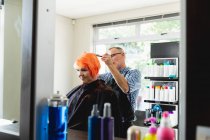 Vista laterale di un parrucchiere caucasico di mezza età e di una giovane donna caucasica che ha i capelli colorati di rosso vivo in un salone di parrucchiere, riflessi in uno specchio — Foto stock