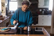 Вид на старших кавказька жінка на кухні наливаючи каву з планшетом поруч з нею і шафи у фоновому режимі — стокове фото