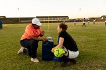 Вид сбоку на тренера смешанной расы средних лет по регби, стоящего на коленях и разговаривающего с молодой белой женщиной-регбисткой на поле для регби во время тренировки — стоковое фото