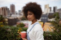 Портрет крупним планом молодої змішаної жінки, що стоїть зовні на балконі в місті, тримає чашку кави і повертає голову, щоб подивитися на камеру — стокове фото