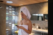 Вид збоку молодий Кавказький жінка брюнетка носіння рушник і з її волосся загорнуті в рушник, використовуючи смартфон у сучасній ванній — стокове фото