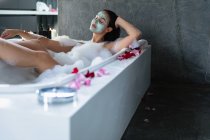 Vista frontal de uma jovem mulher caucasiana vestindo um pacote facial reclinado com os pés para cima em um banho de espuma com pétalas de rosa ao redor da borda — Fotografia de Stock