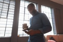 Seitenansicht eines jungen afrikanisch-amerikanischen Mannes, der einen Kaffee zum Mitnehmen in der Hand hält und ein Smartphone vor einem Fenster in einem kreativen Büro benutzt, das von Sonnenlicht hinterleuchtet wird — Stockfoto