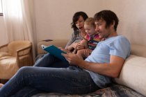 Vista laterale di un giovane padre e madre caucasici seduti su un divano e che leggono un libro con il loro bambino — Foto stock