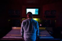 Vue arrière d'un jeune ingénieur du son caucasien debout à un bureau de mixage dans un studio d'enregistrement regardant un moniteur d'ordinateur — Photo de stock