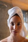 Porträt einer jungen kaukasischen Brünetten mit in ein Handtuch gehüllten Haaren, die in einem modernen Badezimmer in die Kamera lächelt — Stockfoto
