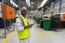 Vue de côté gros plan d'une jeune ouvrière afro-américaine debout à l'aide d'une tablette et souriant à la caméra dans un entrepôt d'une usine de transformation — Photo de stock