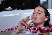 Nahaufnahme einer jungen kaukasischen brünetten Frau, die mit einer brennenden Kerze an der Seite und Rosenblättern darin in einer Badewanne liegt und Champagner mit geschlossenen Augen trinkt — Stockfoto