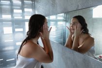 Вид збоку крупним планом молодий Кавказький жінка брюнетка носіння рушник, дивлячись у дзеркало і масажувати її обличчя пальцями в сучасній ванній кімнаті — стокове фото