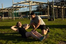 Side view of a young Caucasian woman doing sit ups helped by a young Caucasian man at an outdoor gym during a bootcamp training session — Stock Photo