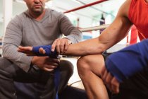 Vorderseite Mittelteil eines jungen kaukasischen männlichen Boxers in einem Boxring, dessen Hände von einem kaukasischen männlichen Trainer mittleren Alters umwickelt werden — Stockfoto