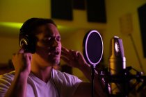 Vista lateral de cerca de un joven cantante de raza mixta con auriculares y cantando frente a un micrófono en un estudio de grabación, con los ojos cerrados - foto de stock