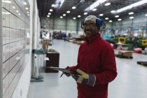 Вигляд збоку закритий середнього віку Афроамериканський робітник на фабриці, який тримає на дошці перевірку на дошці, звертаючись до камери і посміхаючись на складі на заводі обробки. — стокове фото