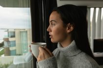 Vista laterale da vicino di una giovane donna bruna caucasica con un maglione a collo alto grigio, che guarda fuori da una finestra con una tazza di caffè, edifici visibili sullo sfondo — Foto stock