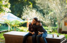 Vue de face de deux pré adolescents garçons caucasiens assis sur un banc dans un jardin, manger le déjeuner et embrasser — Photo de stock