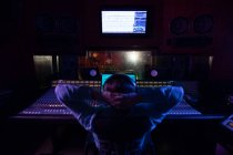 Visão traseira de um jovem engenheiro de som caucasiano sentado com as mãos atrás da cabeça em uma mesa de mistura em um estúdio de gravação assistindo os músicos na sala de gravação através de uma janela — Fotografia de Stock