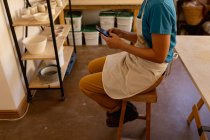 Vista lateral sección baja de alfarera femenina con un delantal sentado en un taburete usando un teléfono inteligente en un estudio de cerámica - foto de stock