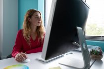 Вигляд збоку закриває молоду кавказьку жінку, яка сидить за столом біля вікна за допомогою комп'ютера в сучасному офісі творчого бізнесу. — стокове фото