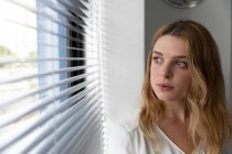 Front view close up of a young Caucasian woman standing with her head turned looking out of a window with venetian blinds in the modern office of a creative business — Stock Photo