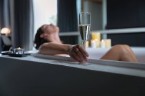 Seitenansicht einer jungen kaukasischen brünetten Frau, die mit brennenden Kerzen in einem Bad liegt, sich zurücklehnt und ein Glas Champagner in der Hand hält — Stockfoto
