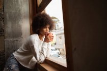 Vue de côté gros plan d'une jeune femme métisse penchée sur un rebord de fenêtre regardant dehors boire une tasse de café — Photo de stock