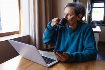 Vista frontal de una mujer caucásica mayor sentada en una mesa en casa bebiendo café y sosteniendo un teléfono inteligente, con una computadora portátil frente a ella - foto de stock