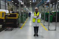 Retrato de uma mulher caucasiana de meia-idade vestindo óculos e roupas de trabalho entre fileiras de equipamentos sorrindo em um armazém em uma fábrica de processamento, outro trabalhador visível no fundo — Fotografia de Stock