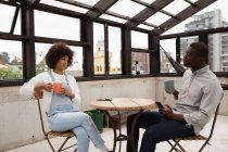 Vista lateral de una joven mestiza y un joven afroamericano sentados en una mesa tomando café y hablando en una habitación cubierta de cristal en una azotea, con edificios de la ciudad en el fondo . - foto de stock