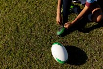 High angle low section of female rugby player sitting and tying her boot on a rugby pitch, with the ball beside her — Stock Photo
