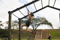 Side view of a young Caucasian woman hanging from monkey bars at an outdoor gym during a bootcamp training session, with another participant in the background — Stock Photo