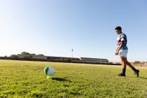 Vista lateral de una joven jugadora de rugby de raza mixta adulta parada en un campo de rugby preparándose para correr hasta la pelota y hacer una patada en el lugar - foto de stock